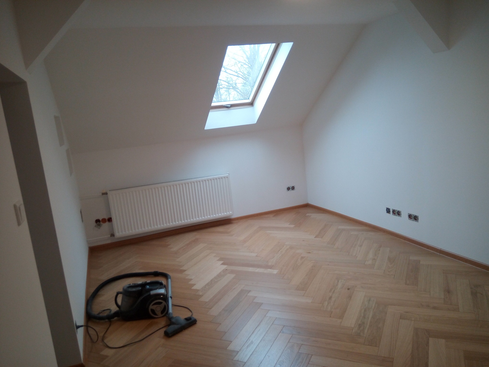 Poddasze po remoncie z parkietem ułożonym w jodełkę, grzejnikiem i oknem dachowym, z odkurzaczem w rogu.