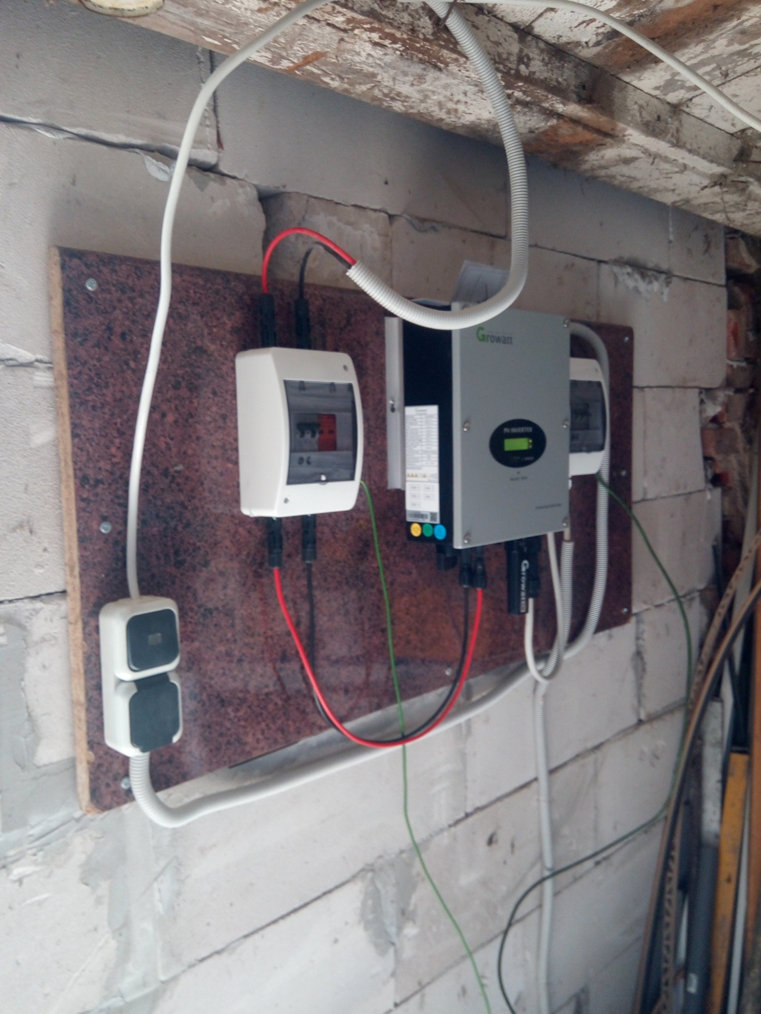 Szary inwerter Growatt z wyświetlaczem, biała skrzynka bezpiecznikowa i podwójne gniazdko zamontowane na brązowej płycie na ścianie z białych bloczków, widoczne kable i rurki instalacyjne.