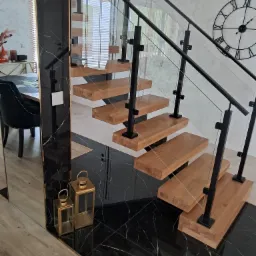 Nowoczesne schody metalowe z drewnianymi stopniami i szklanymi balustradami, czarne elementy konstrukcyjne, marmurowe wykończenie podłogi i ściany, zegar na ścianie w tle.