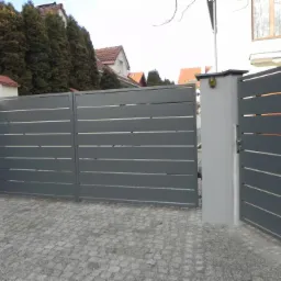 Nowoczesna, szara brama wjazdowa z poziomymi panelami, częściowo otwarta, z widocznym mechanizmem zamka i kostką brukową na podjeździe.