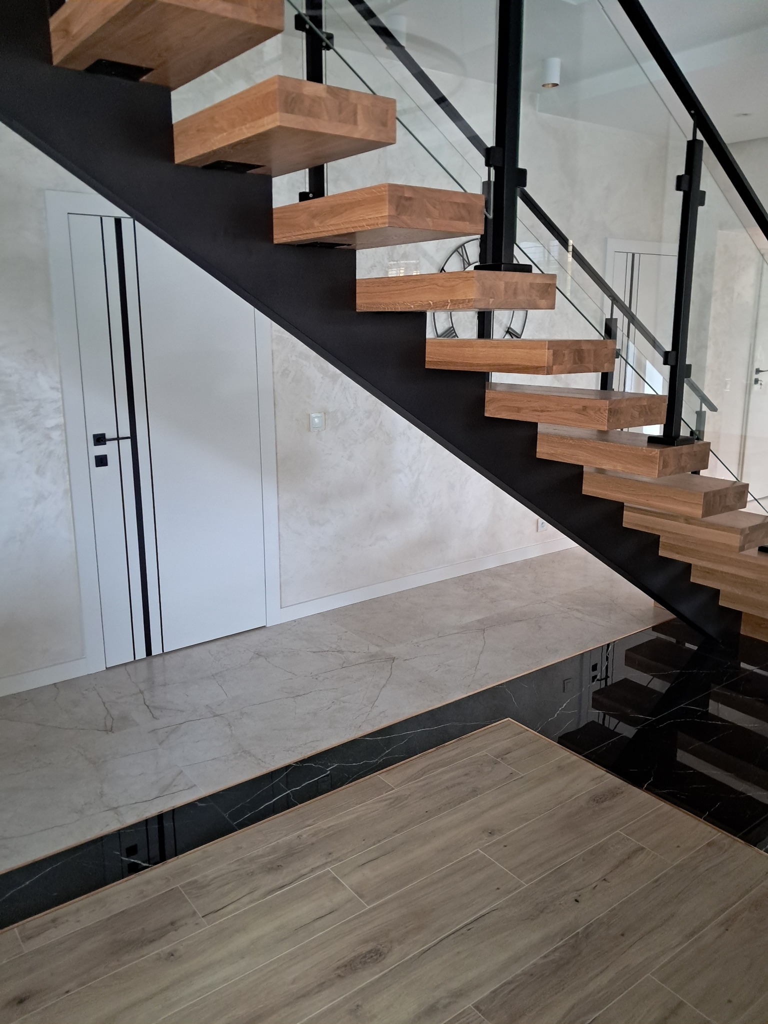 Nowoczesne schody z drewnianymi stopniami i czarną metalową konstrukcją oraz szklaną balustradą, widoczne także drzwi z czarnymi paskami i różnorodne płytki podłogowe.