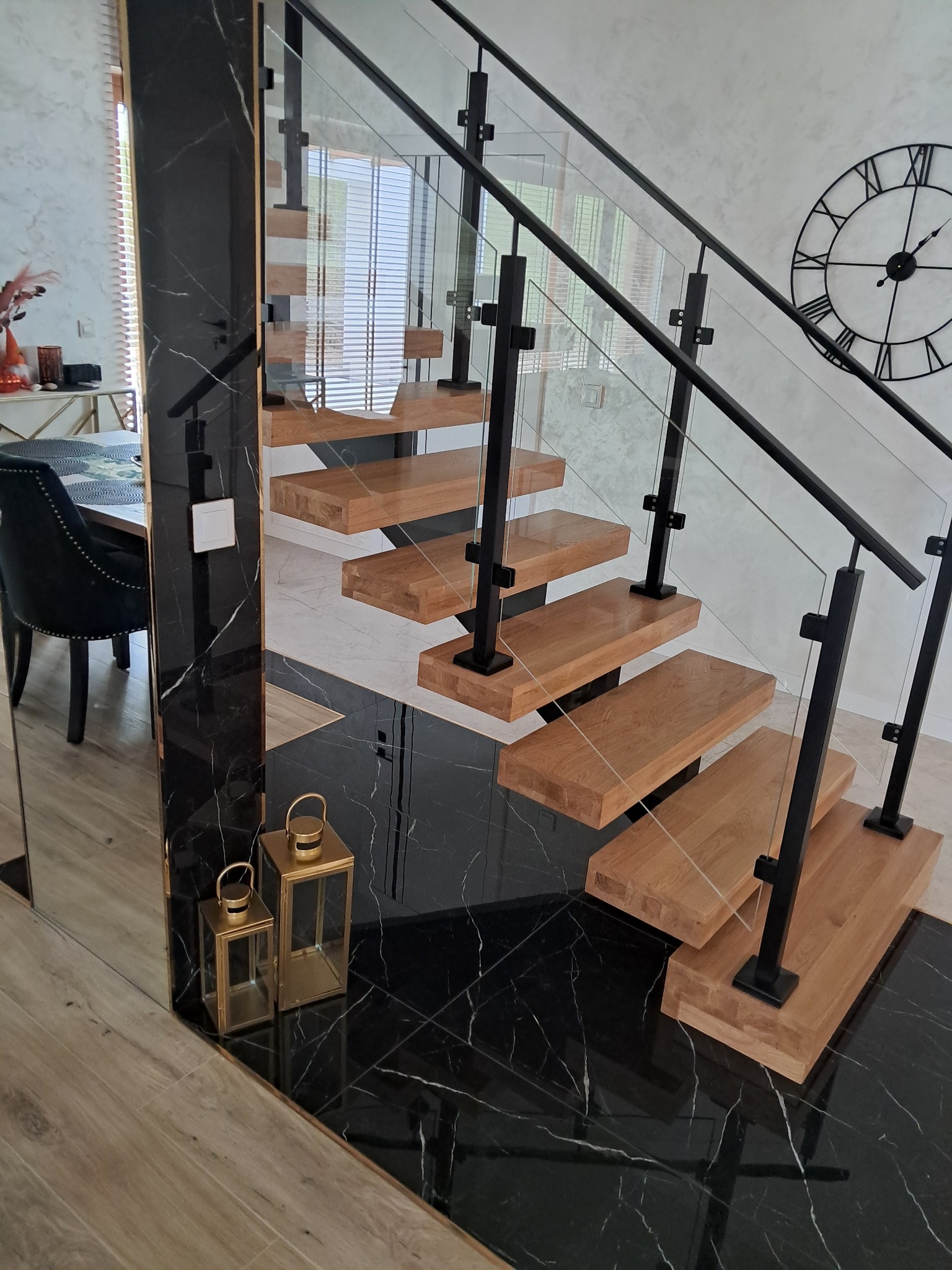 Nowoczesne schody metalowe z drewnianymi stopniami i szklanymi balustradami, czarne elementy konstrukcyjne, marmurowe wykończenie podłogi i ściany, zegar na ścianie w tle.