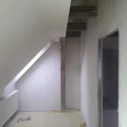 Poddasze w trakcie adaptacji: ściany wykończone gładzią, widoczna drewniana belka konstrukcyjna, grzejnik i zabezpieczona folią podłoga, przygotowany otwór drzwiowy.