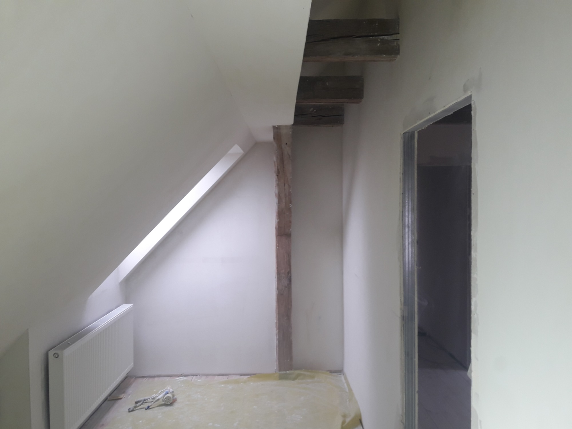 Poddasze w trakcie adaptacji: ściany wykończone gładzią, widoczna drewniana belka konstrukcyjna, grzejnik i zabezpieczona folią podłoga, przygotowany otwór drzwiowy.