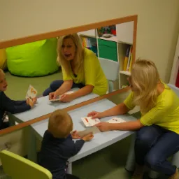 Logopeda prowadzi zajęcia z chłopcem przy stole, odbicie w lustrze ukazuje dodatkowy widok na interakcję, w tle regał z książkami i zielona pufa.