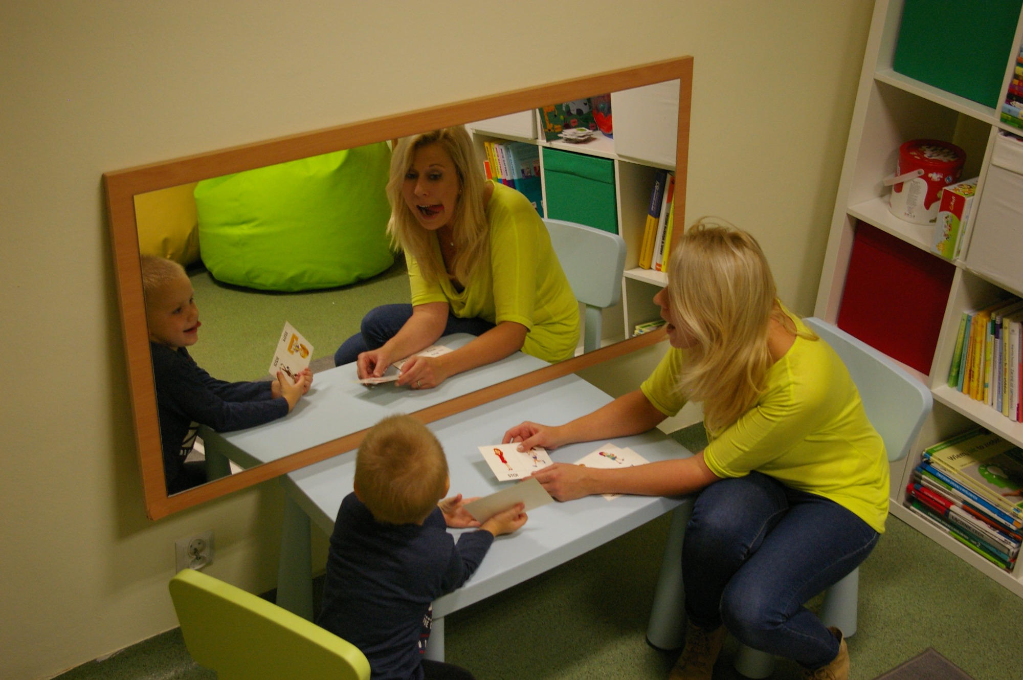 Logopeda prowadzi zajęcia z chłopcem przy stole, odbicie w lustrze ukazuje dodatkowy widok na interakcję, w tle regał z książkami i zielona pufa.
