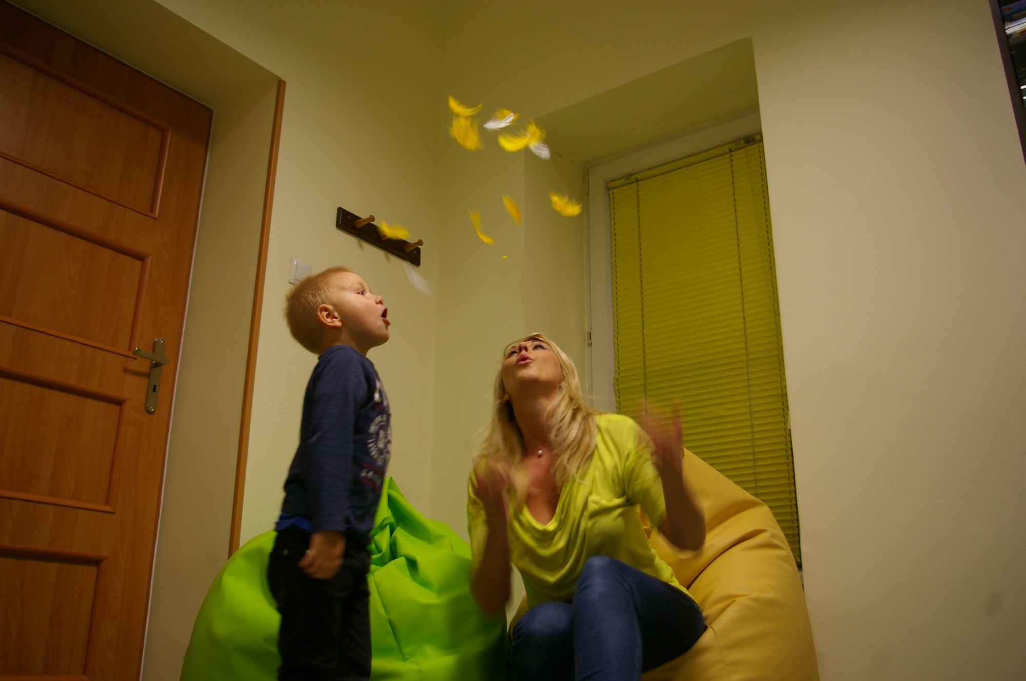 Kobieta rzuca w górę piórka, na które patrzy z otwartymi ustami mały chłopiec stojący obok zielonej pufy.