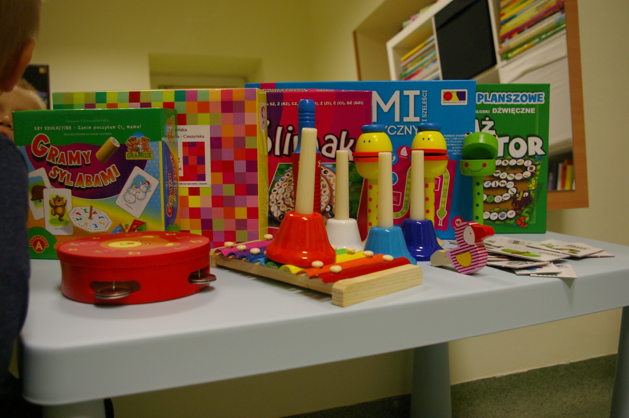 Kolorowe gry edukacyjne i instrumenty muzyczne dla dzieci na niebieskim stole, w tle regał z książkami.