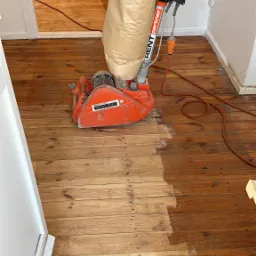 Szlifowanie drewnianej podłogi parkieciarką; widoczna różnica w kolorze między obszarem wyszlifowanym a nieobrobionym, w tle widoczna kolejna maszyna.
