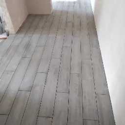 Ułożona podłoga z szarych, podłużnych płytek imitujących drewno, widok z perspektywy wejścia do pomieszczenia z surowymi ścianami w trakcie wykończenia.