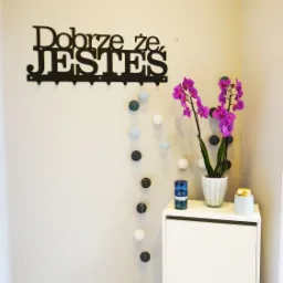 Jasne wnętrze z białymi meblami, fioletową orchideą w doniczce, dekoracyjnym napisem 'Dobrze że JESTEŚ' na ścianie i girlandą z kul.