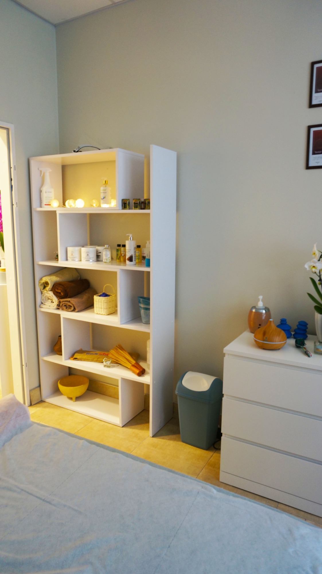 Jasne wnętrze gabinetu z łóżkiem przykrytym prześcieradłem, obok biały regał z kosmetykami, ręcznikami i akcesoriami oraz biała komoda z dyfuzorem zapachowym i kwiatami.