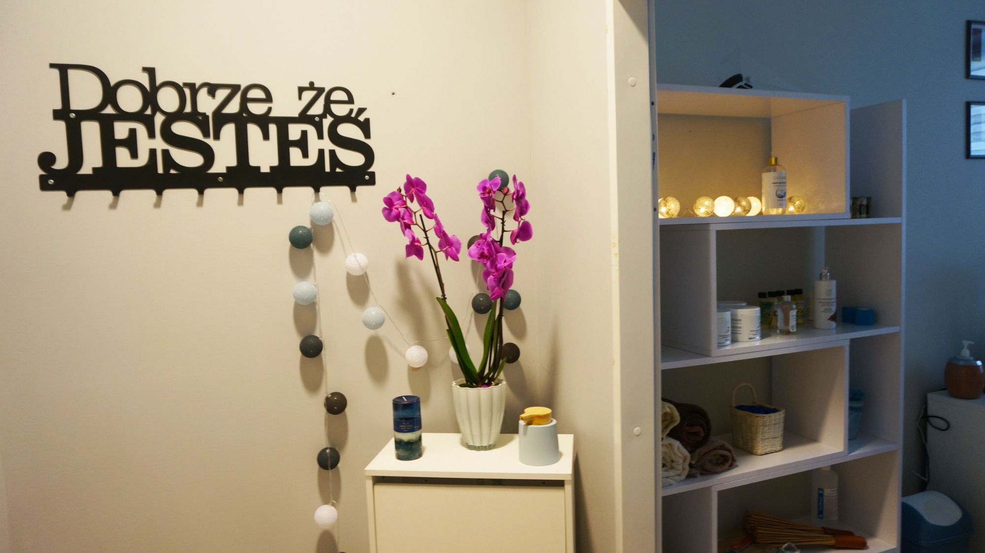 Jasny gabinet z dekoracjami: fioletowe orchidee w białej doniczce, świeca, dozownik na białej szafce, napis 'Dobrze że jesteś' z haczykami, girlanda z kulkami, biały regał z kosmetykami i ręcznikami.