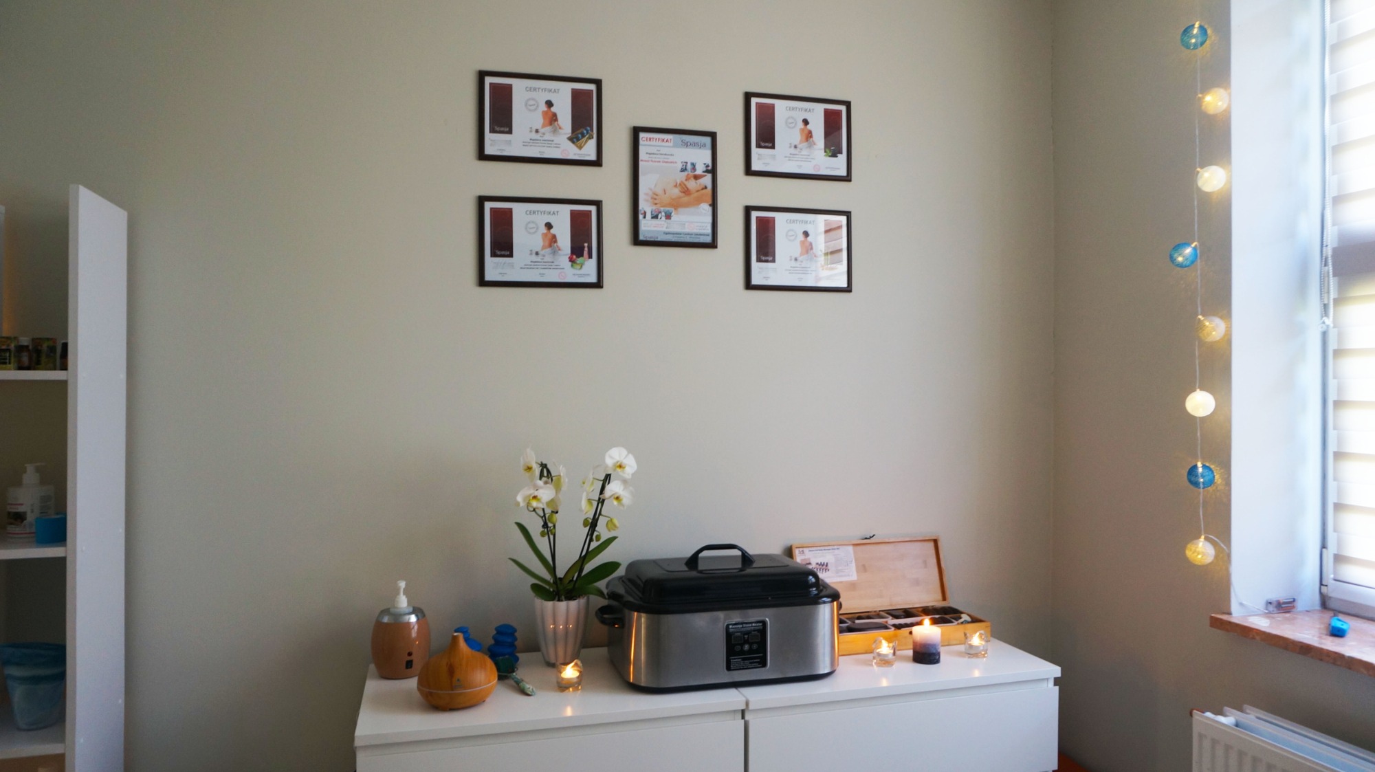 Gabinet z certyfikatami na ścianie, szafką z podgrzewaczem do kamieni, dyfuzorem zapachowym, świecami i storczykiem.