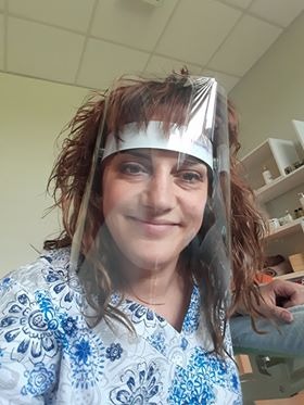 Uśmiechnięta kobieta w bluzie w kwiaty i przezroczystej masce ochronnej na twarzy, prawdopodobnie fizjoterapeutka lub masażystka w gabinecie.