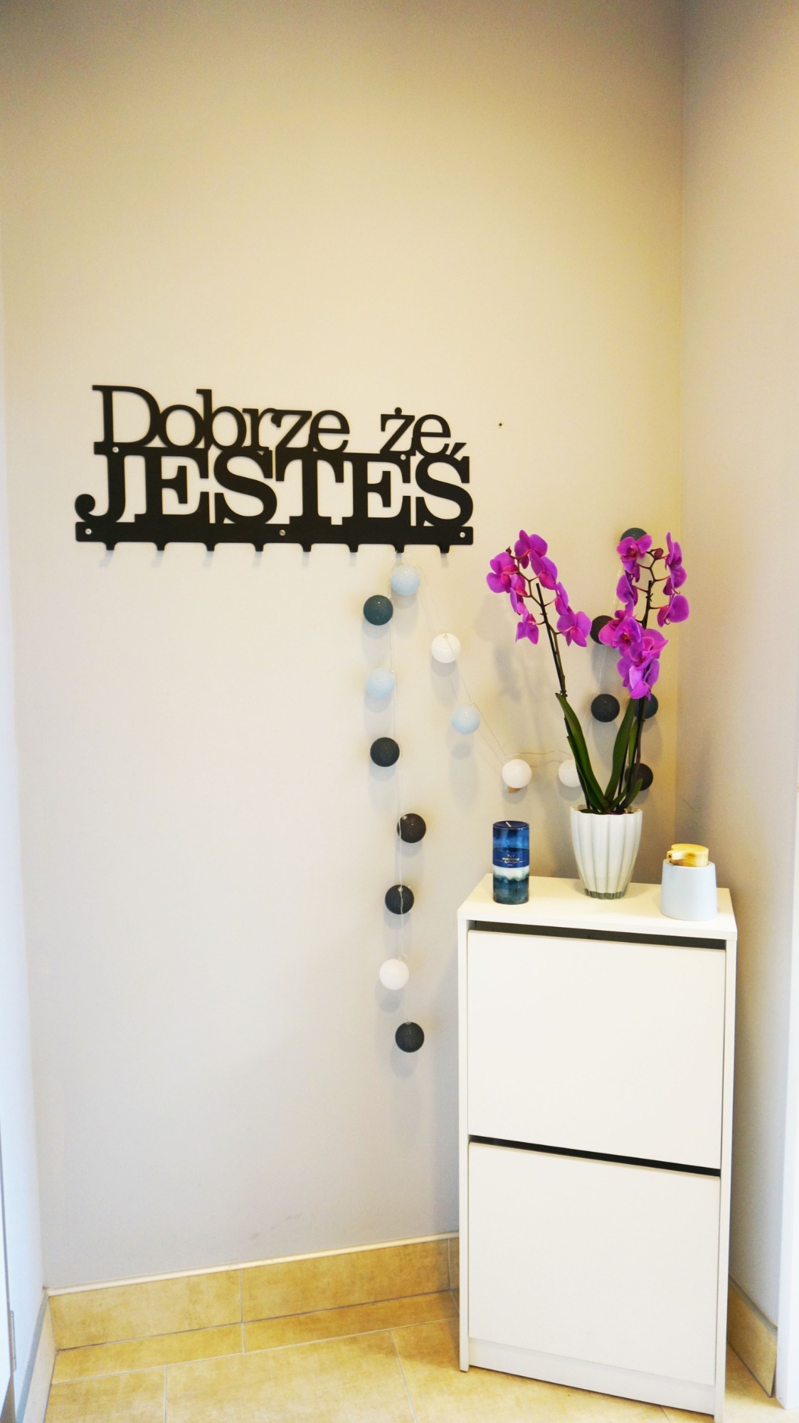 Jasne wnętrze z białymi meblami, fioletową orchideą w doniczce, dekoracyjnym napisem 'Dobrze że JESTEŚ' na ścianie i girlandą z kul.