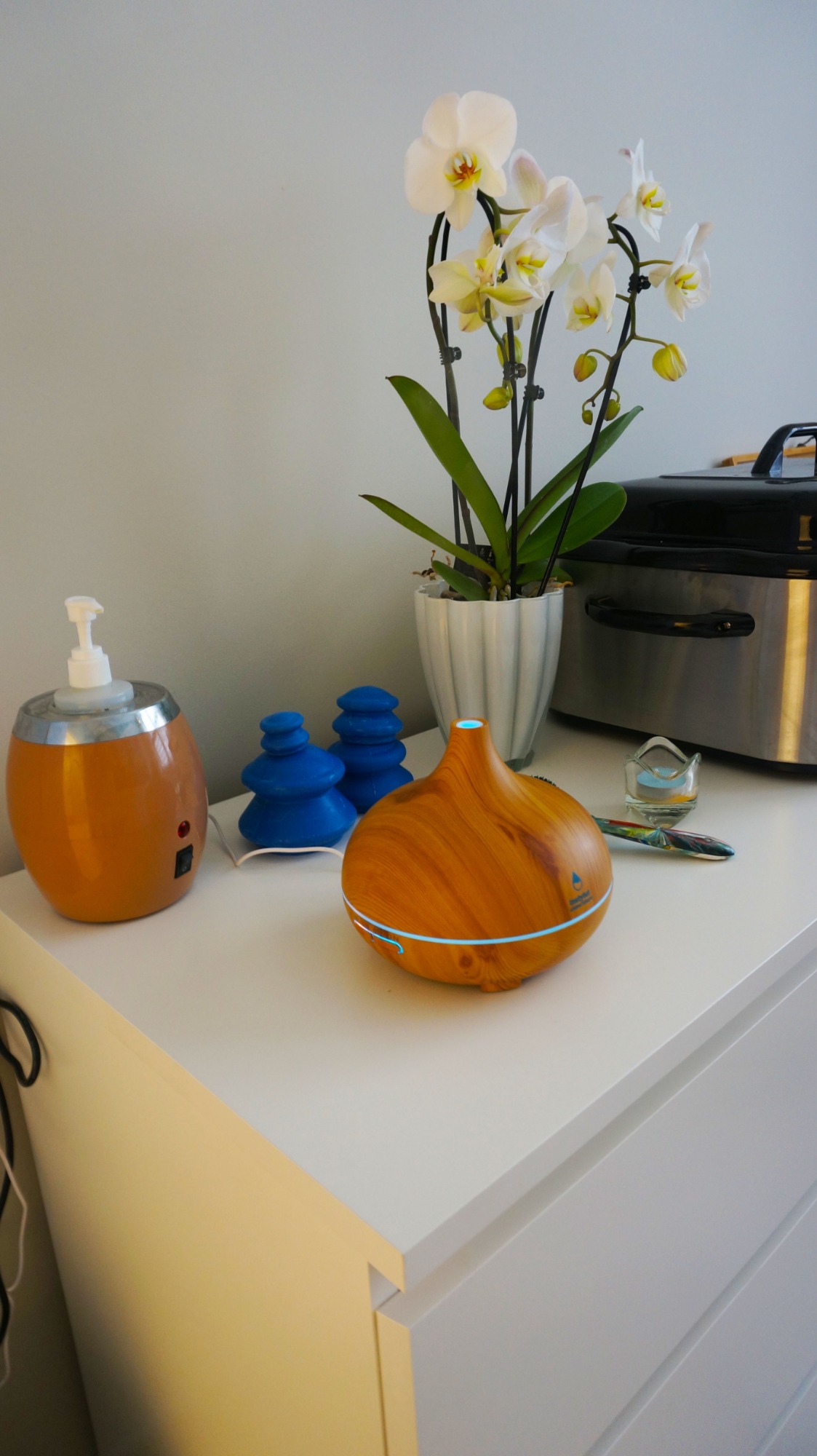 Aromaterapia: dyfuzor zapachowy z efektem drewna, podgrzewacz do olejków, kamienie do masażu i storczyk na białej komodzie.