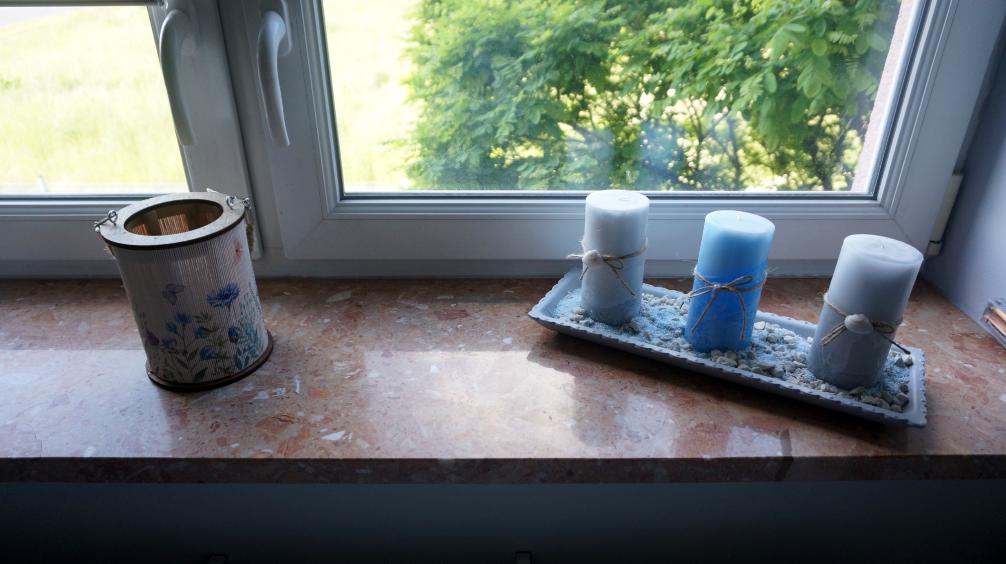 Dekoracja okienna z trzema świecami w odcieniach bieli i błękitu na ozdobnej tacy z drobnymi kamyczkami oraz lampion z motywem kwiatowym, ustawione na marmurowym parapecie przy oknie z widokiem...