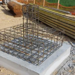 Zbrojenie fundamentu na betonowej podstawie, widoczne deski szalunkowe i siatka zabezpieczająca w tle.