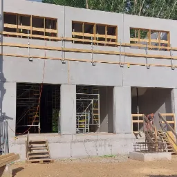 Wznoszenie dwukondygnacyjnego, surowego budynku z prefabrykat&oacute;w żelbetowych na placu budowy, z ż&oacute;łtym dźwigiem w tle i widocznymi rusztowaniami oraz pracownikiem wchodzącym po schodach.