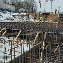 Widok na plac budowy z przygotowanym zbrojeniem fundamentów, deskowaniem i niwelatorem laserowym na trójnogu, w tle pracownicy i ciężki sprzęt budowlany.