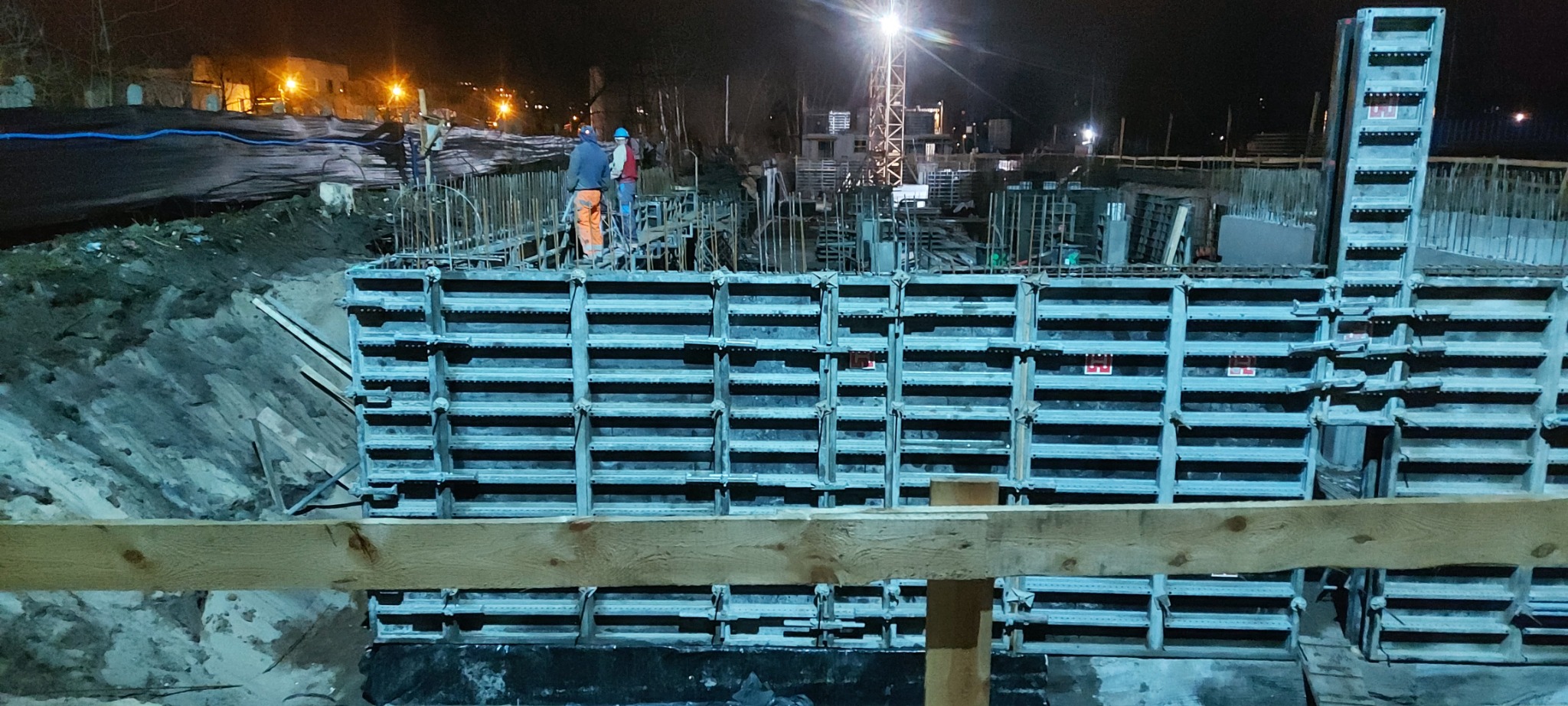 Nocna budowa: szalunek pod fundamenty z widocznym zbrojeniem i pracownikami w oddali, oświetlona lampami budowlanymi.