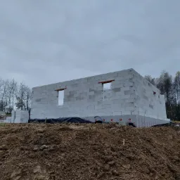 Wznoszenie ścian parterowego budynku z betonu komórkowego na nierównym terenie, widoczne otwory okienne i przygotowane bloczki, pracownik w tle.