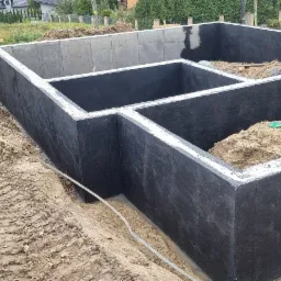 Zabezpieczone hydroizolacją betonowe fundamenty domu jednorodzinnego, widoczne ślady opon koparki na ziemi.