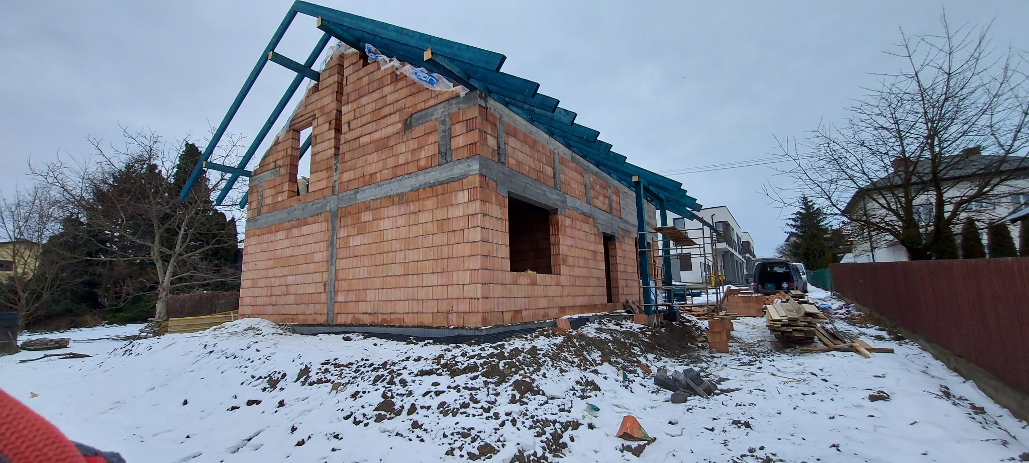 Budowa domu z czerwonej cegły w zimowej scenerii, widoczne niebieskie elementy konstrukcji dachu i częściowo ośnieżone otoczenie.