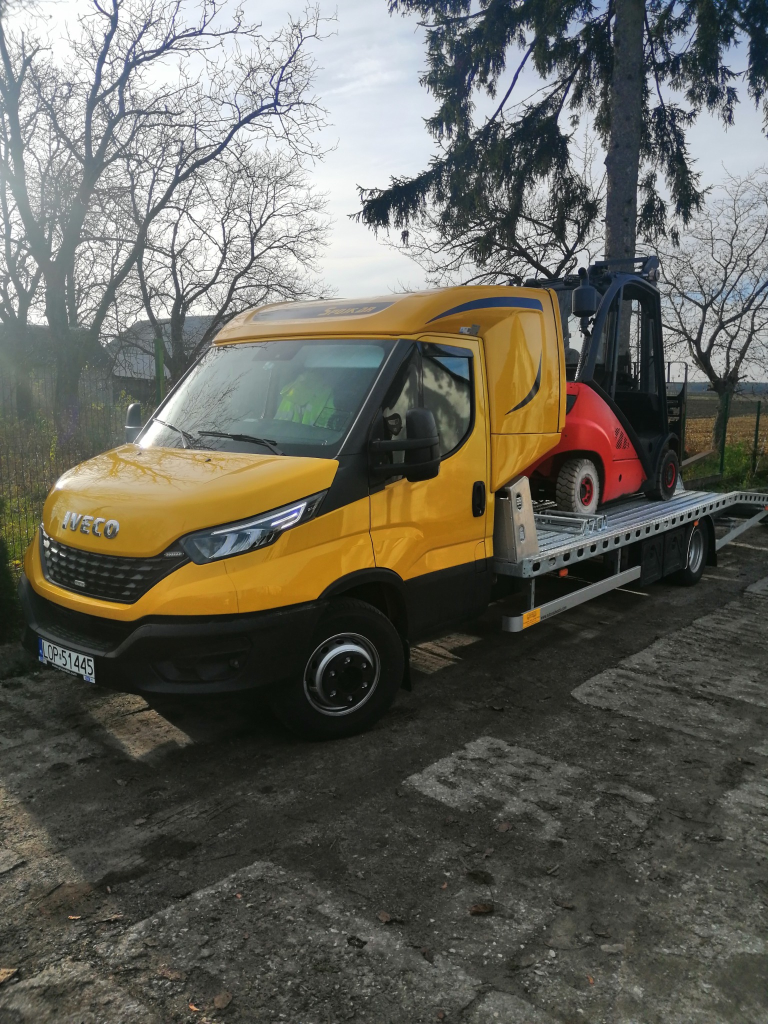 Żółto-czarny Iveco na platformie transportowej przewozi czerwony wózek widłowy, na tle drzew bez liści i częściowo pochmurnego nieba.