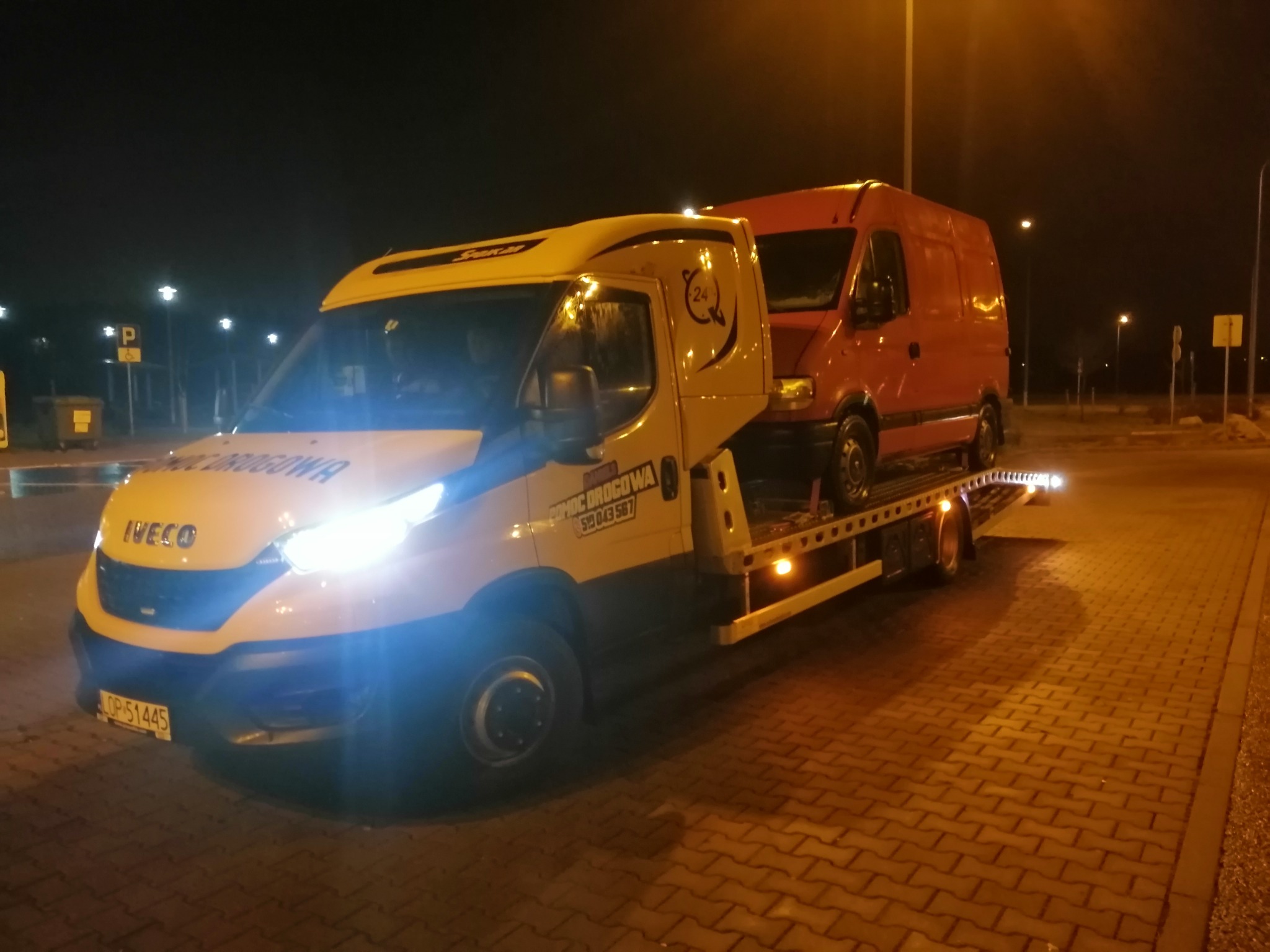 Żółta ciężarówka pomocy drogowej Iveco z pomarańczowym vanem na platformie, sfotografowana nocą na parkingu z kostki brukowej, z włączonymi światłami. Widoczny numer telefonu kontaktowego.