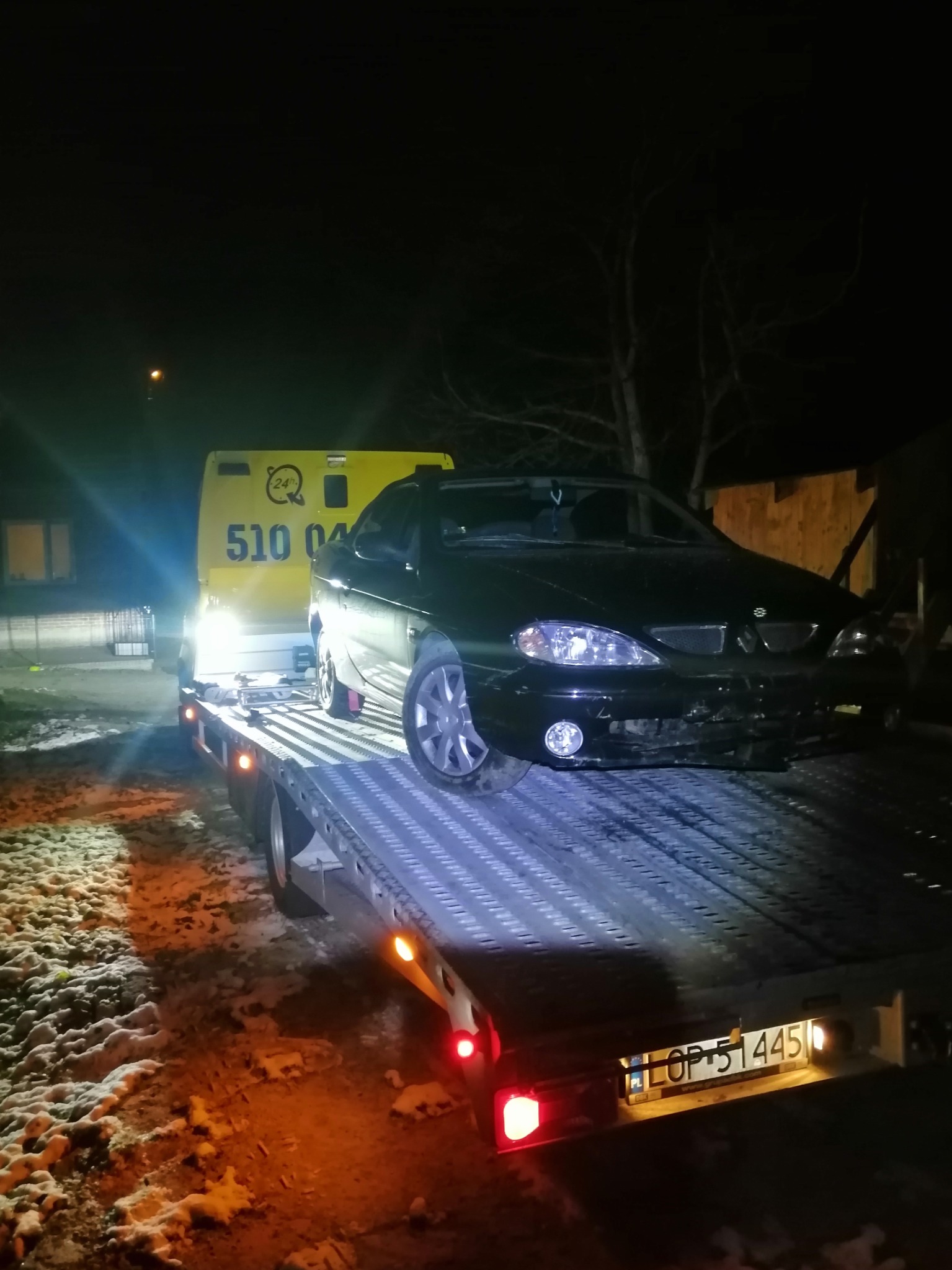 Czarny samochód osobowy z uszkodzonym przodem załadowany na platformę pojazdu pomocy drogowej w nocy, widoczny numer telefonu na żółtej kabinie