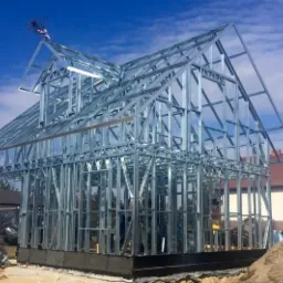 Konstrukcja szkieletowa domu w trakcie budowy, widoczne stalowe profile, fundamenty i pracownicy budowlani w tle.