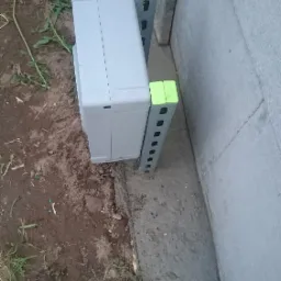 Szara skrzynka telekomunikacyjna na ziemi, wsparta na dw&oacute;ch metalowych wspornikach z jaskrawo zielonymi nakładkami, przy betonowym fundamencie i ścianie.