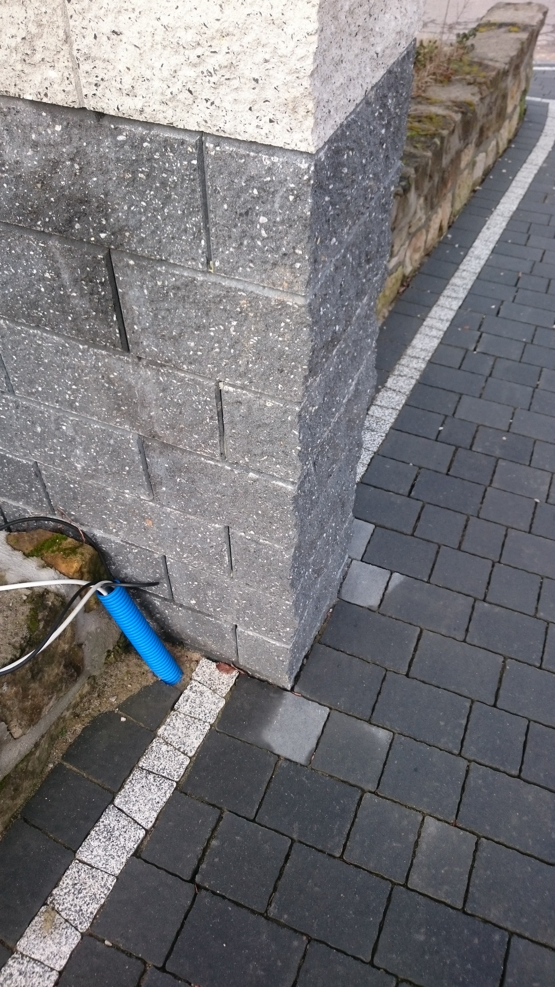 Szara, murowana kolumna z widoczną niebieską rurą karbowaną i przewodami wychodzącymi z ziemi, obok ułożona kostka brukowa.