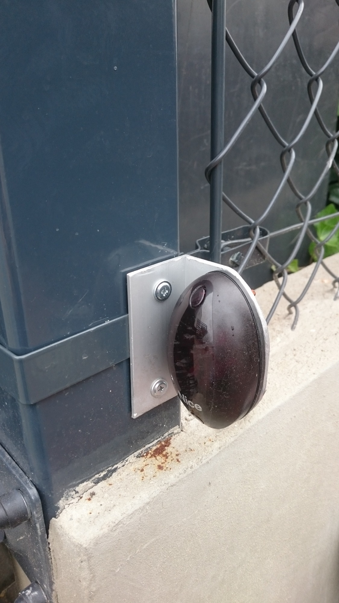 Fotokomórka marki Nice zamontowana na słupku bramy w Wierzbicy, widoczne śruby mocujące i fragment siatki ogrodzeniowej w tle.