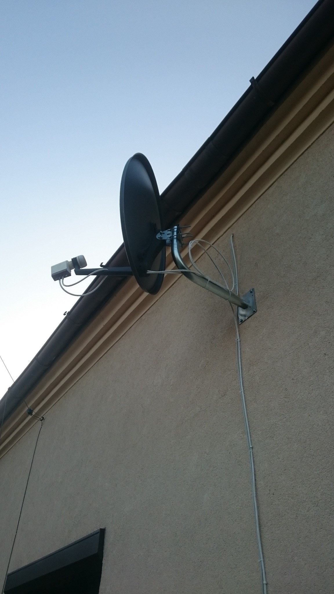 Czarna antena satelitarna zamontowana na ścianie budynku pod rynną, widoczne kable i uchwyt mocujący.