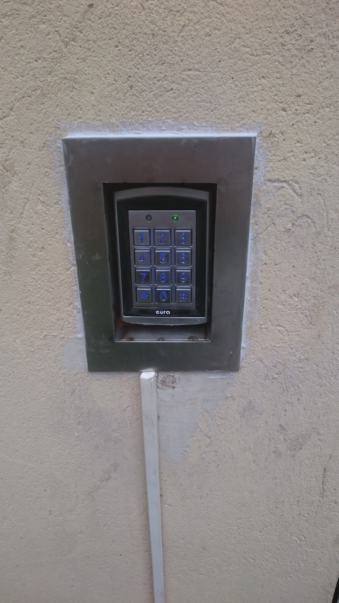 Zewnętrzny panel numeryczny domofonu z podświetlanymi przyciskami, zamontowany w metalowej ramce na szorstkiej ścianie budynku. Widoczny biały kanał kablowy.