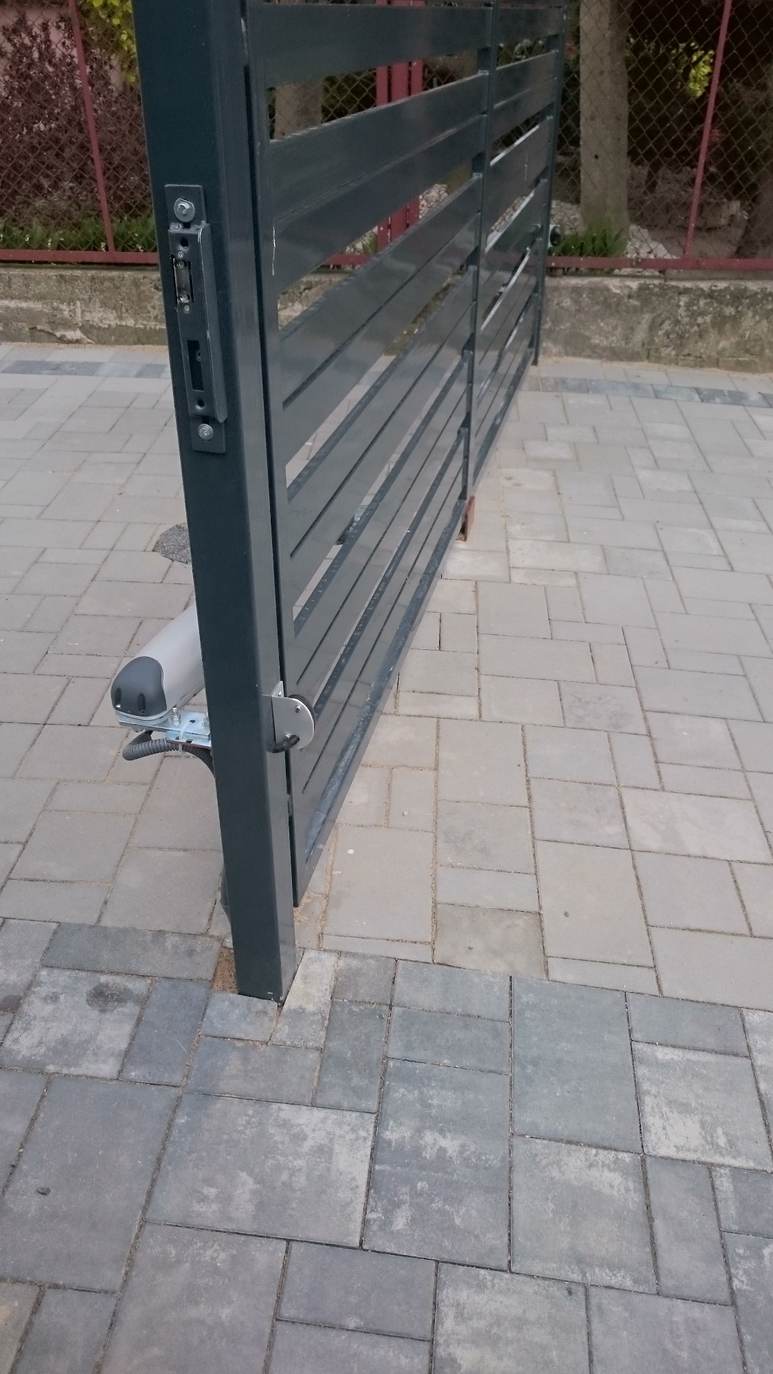 Szara, metalowa brama wjazdowa z automatycznym napędem, widoczna na tle kostki brukowej w Wierzbicy. Brama częściowo otwarta, z widocznym mechanizmem zamka i siłownikiem.