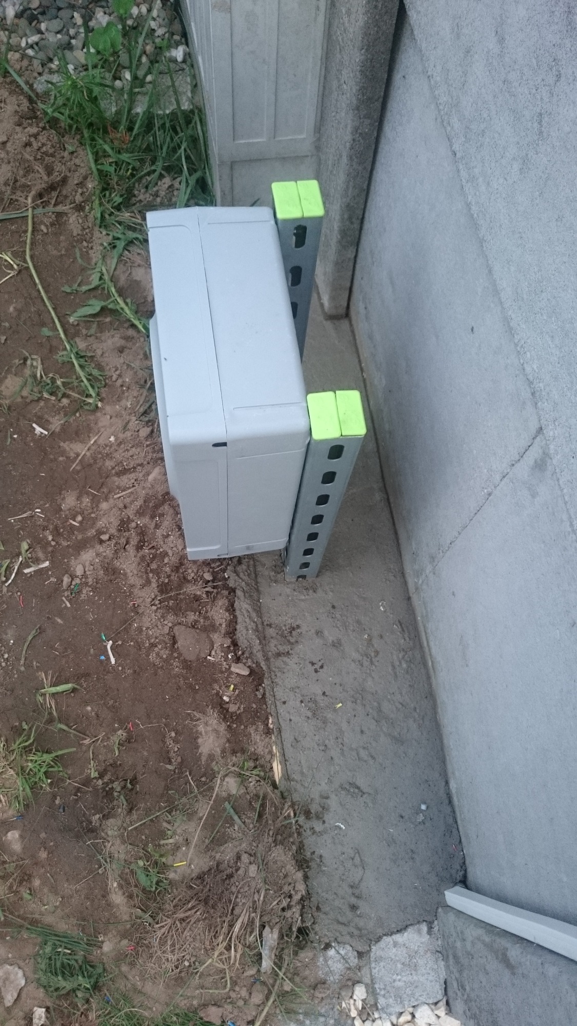 Szara skrzynka telekomunikacyjna na ziemi, wsparta na dwóch metalowych wspornikach z jaskrawo zielonymi nakładkami, przy betonowym fundamencie i ścianie.