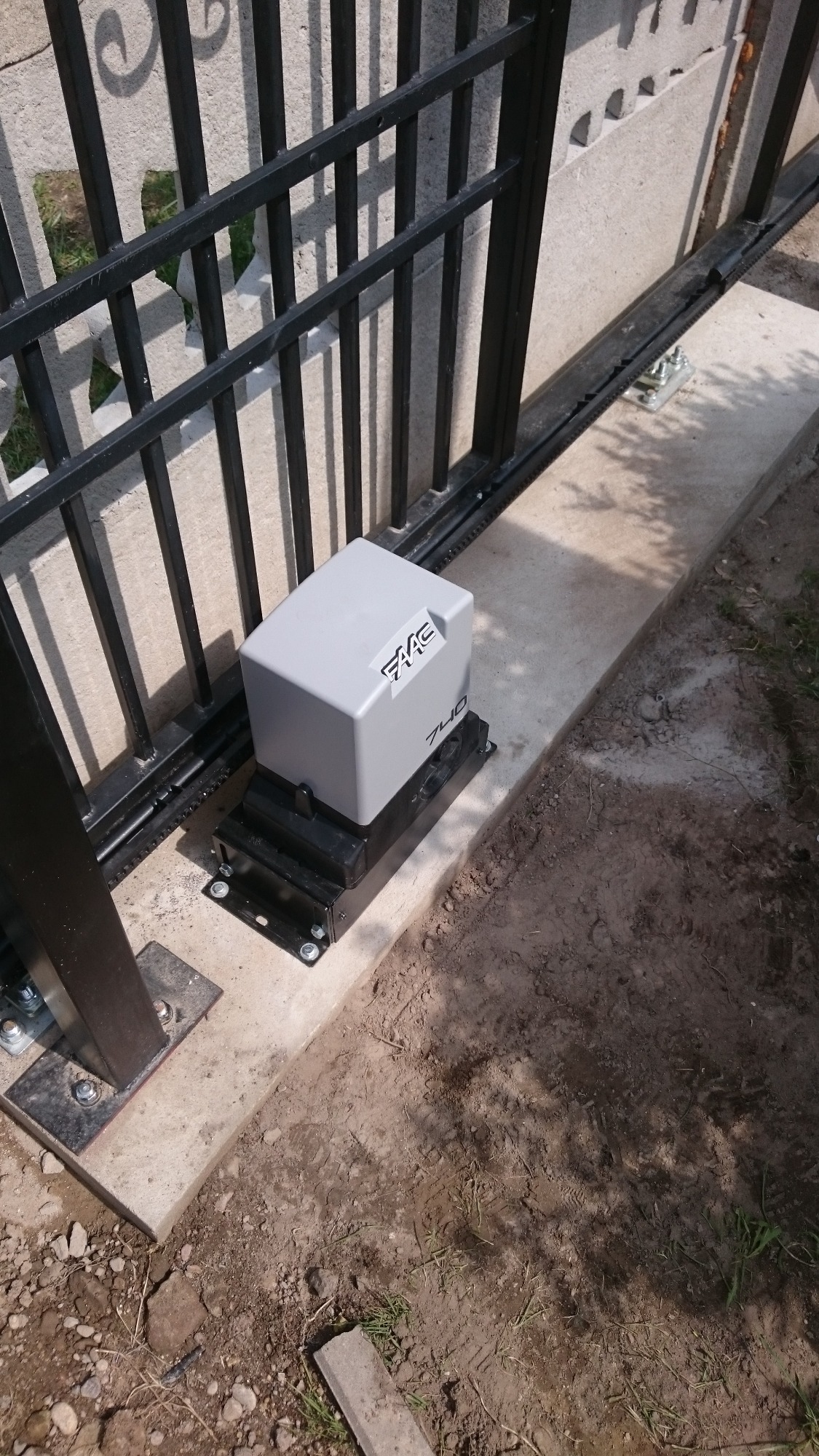 Szary napęd bramy przesuwnej marki FAAC 740 zamontowany na betonowej podstawie obok metalowej bramy z czarnymi prętami, widoczna listwa zębata.