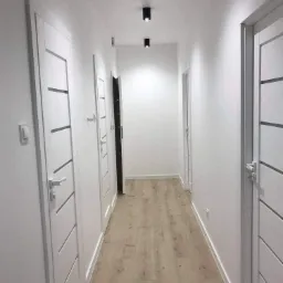Wnętrze korytarza po odświeżeniu, białe ściany, jasna podłoga, trzy białe drzwi z poziomymi wstawkami, czarne lampy sufitowe.
