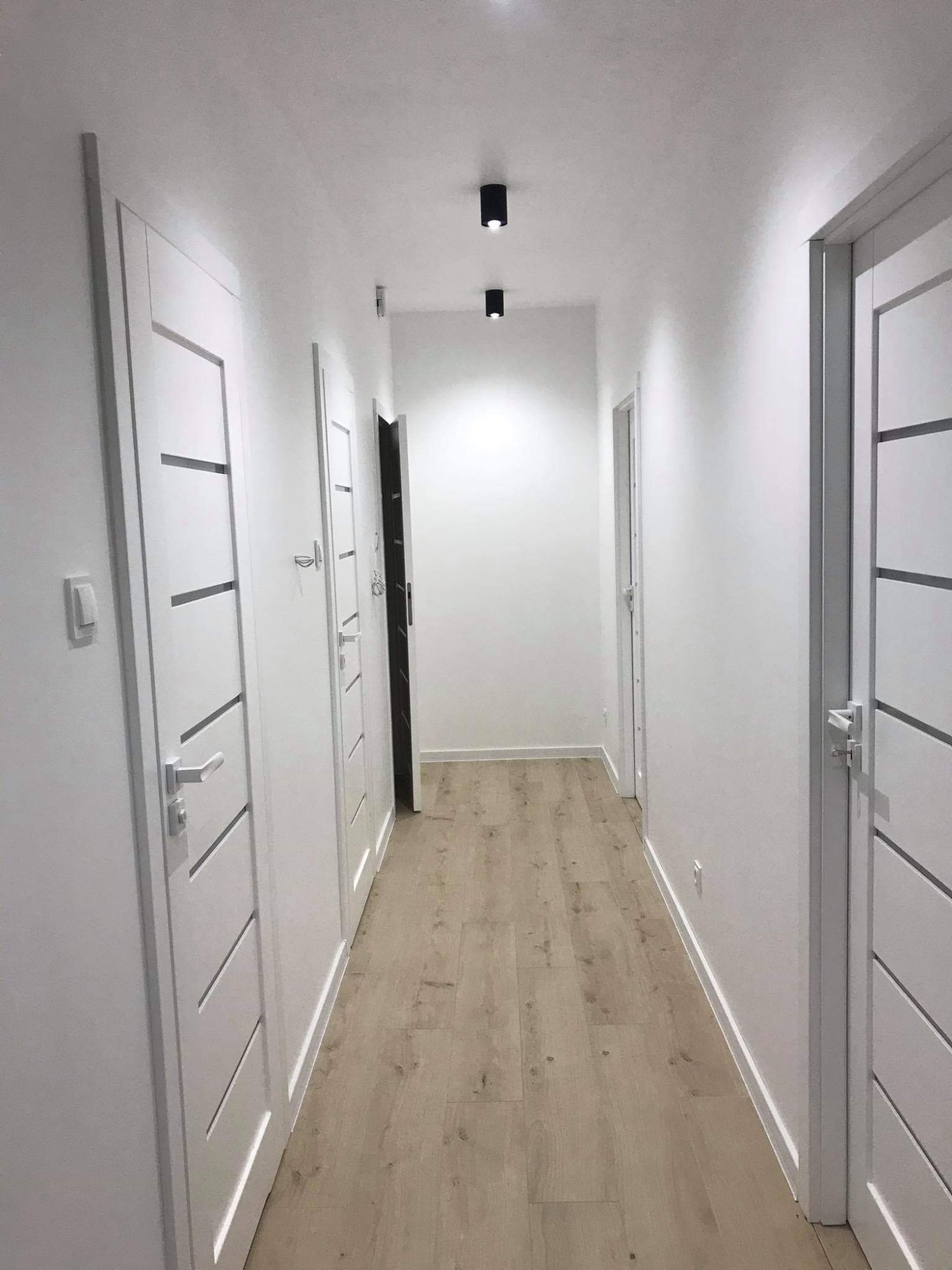 Wnętrze korytarza po odświeżeniu, białe ściany, jasna podłoga, trzy białe drzwi z poziomymi wstawkami, czarne lampy sufitowe.
