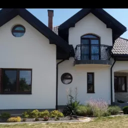 Elewacja domu jednorodzinnego z białym tynkiem, ciemnobrązową stolarką okienną i dachem, z okrągłymi oknami i balkonem z kutą balustradą, otoczona zielenią.