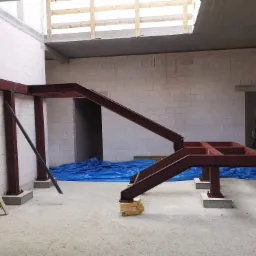 Surowa, stalowa konstrukcja schodów w trakcie montażu w budynku w stanie surowym, zabezpieczona niebieską folią, widoczne drewniane podkładki poziomujące.