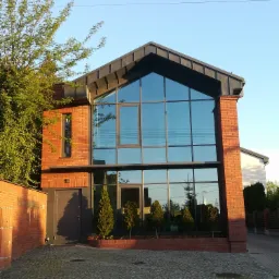 Nowoczesny dom z cegły z dużymi oknami i metalowym dachem w kształcie litery A, otoczony zielenią i brukowanym podjazdem.