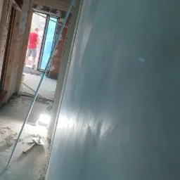 Świeżo nałożony tynk na ścianie w trakcie remontu, widoczny profil narożny, narzędzia na podłodze i sylwetka osoby w oddali przy oknie.