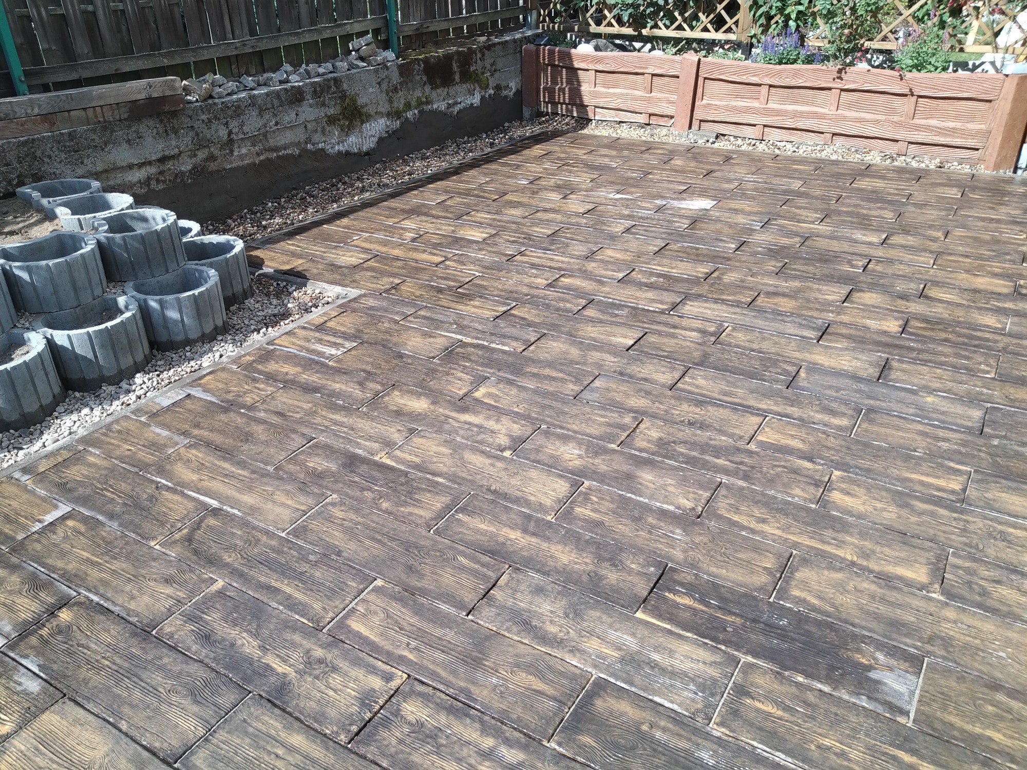 Ułożona kostka brukowa imitująca drewno na podwórku, widoczne betonowe obrzeża i przygotowane miejsce na nasadzenia roślin.