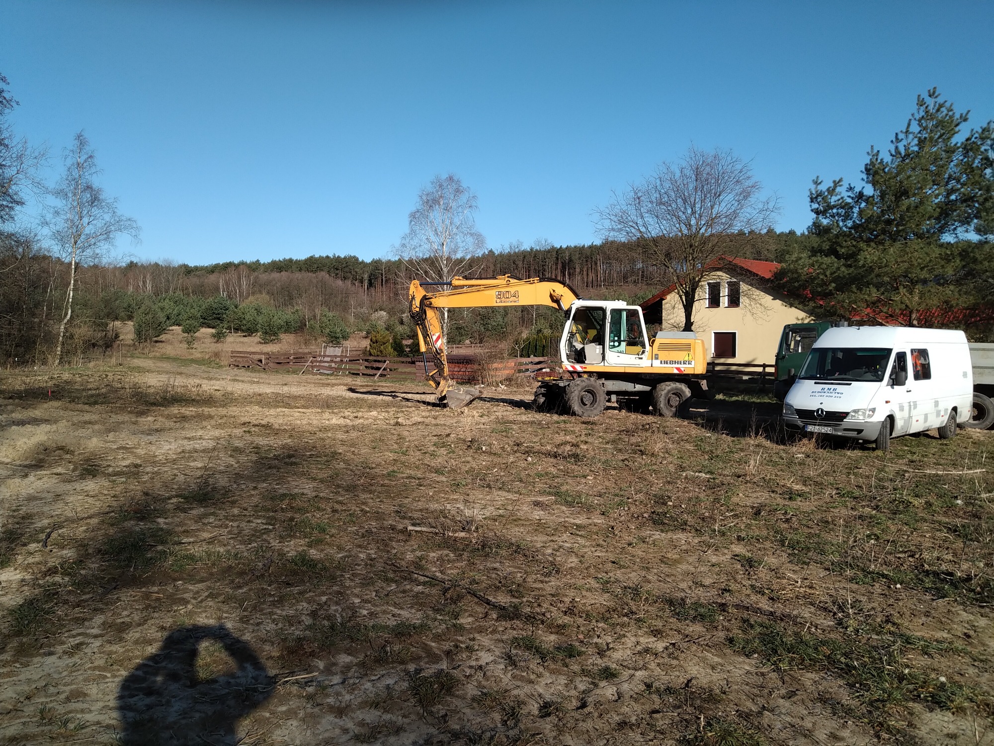 Żółta koparka Liebherr 904, biały van Mercedes-Benz z logo firmy budowlanej na tle domu z czerwonym dachem i drzew w słoneczny dzień. Cień osoby w dolnej części kadru.