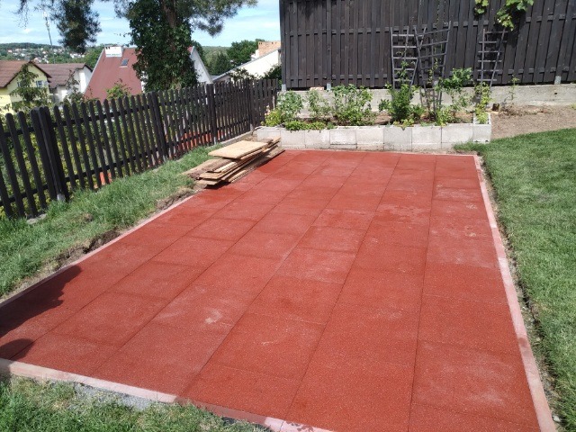 Czerwona, gumowa nawierzchnia ułożona z kwadratowych płyt na przygotowanym podłożu w ogrodzie, widoczne obrzeża i paleta drewnianych desek obok trawnika.